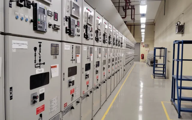 Șir de panouri electrice într-o încăpere industrială cu podea marcată cu linii galbene