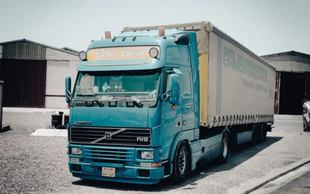 Camion Volvo albastru cu semiremorcă, în fața unor depozite
