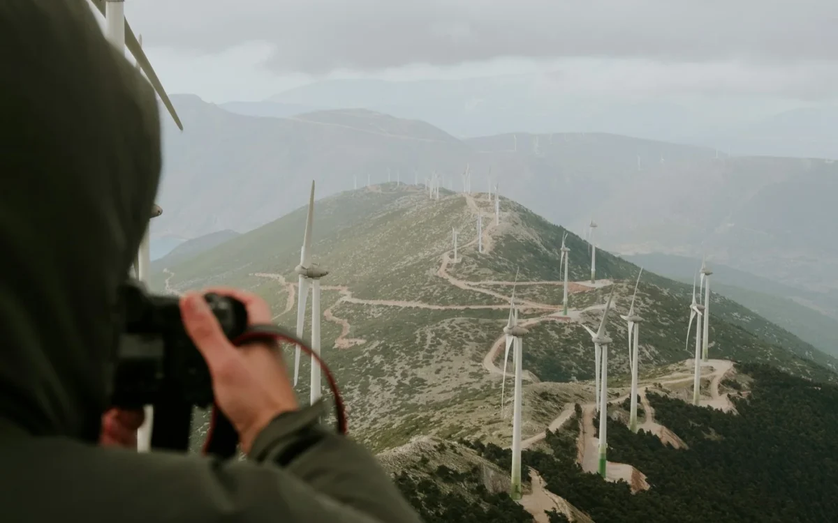 Persoană cu aparat foto în fața unor turbine eoliene amplasate pe coline